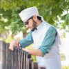A man in white hat and glasses standing next to a fence.